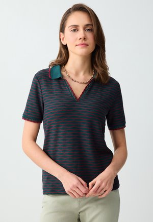 Femme portant un polo à manches courtes rayé vert et rouge avec des bords festonnés et un collier de perles, debout devant un fond uni.