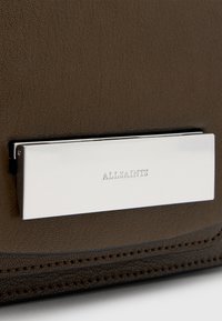 Brun struktureret lædertaske med en metallisk sølvplade prægede med "ALLSAINTS," sikret med en minimalistisk lukkemekanisme.