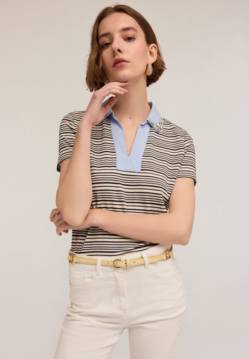 Cămașă polo stripedă în alb și negru, cu un guler bleu deschis, având un design cu decupaj și broderii pe umăr. Purtați-o cu pantaloni albi.