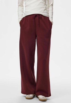 Personne portant un pantalon de survêtement ample rouge foncé avec cordon de serrage à la taille, mains dans les poches, haut blanc à manches longues et baskets blanches avec semelles en gomme.
