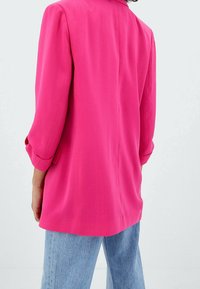 Persona vistiendo un blazer rosa brillante con las mangas arremangadas y jeans de color azul claro, de pie contra un fondo blanco liso.