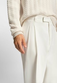 Maglione crema con un motivo a spina di pesce testurizzato; pantaloni bianchi plissettati con una finitura liscia e una cintura piatta, mano appoggiata di lato.