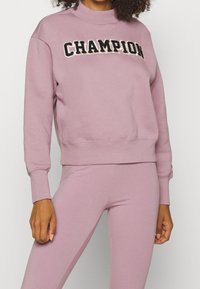 Felpa rosa chiaro con collare e polsini a coste, con la scritta "CHAMPION" ricamata in bianco e nero sul petto.