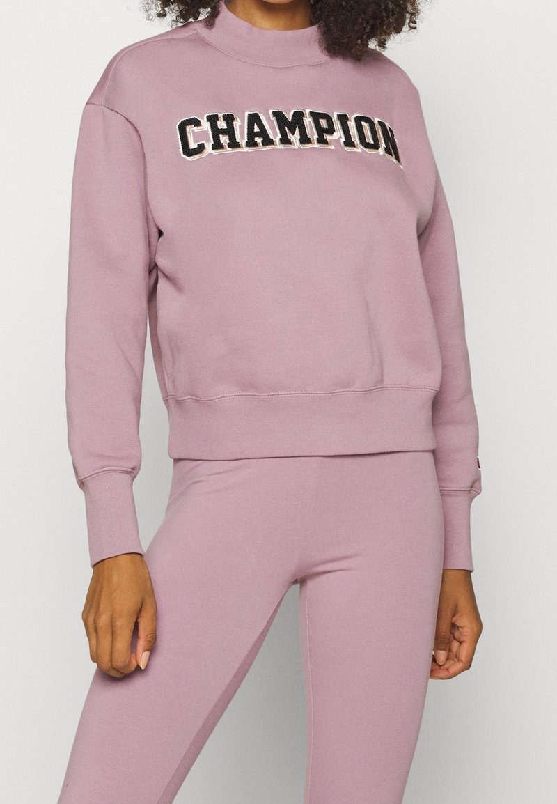 Felpa rosa chiaro con collare e polsini a coste, con la scritta "CHAMPION" ricamata in bianco e nero sul petto.