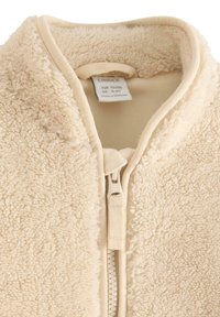 Beige fleecejas met een zachte textuur, hoge kraag en een rits aan de voorkant. Voering met zichtbaar zorglabel. Heeft een consistent stofpatroon.