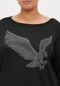 Schwarzes Langarmshirt mit einem großen silbernen, mit Nieten besetzten Adler-Graphic auf der Vorderseite, abgerundetem Halsausschnitt und geripptem Textur.
