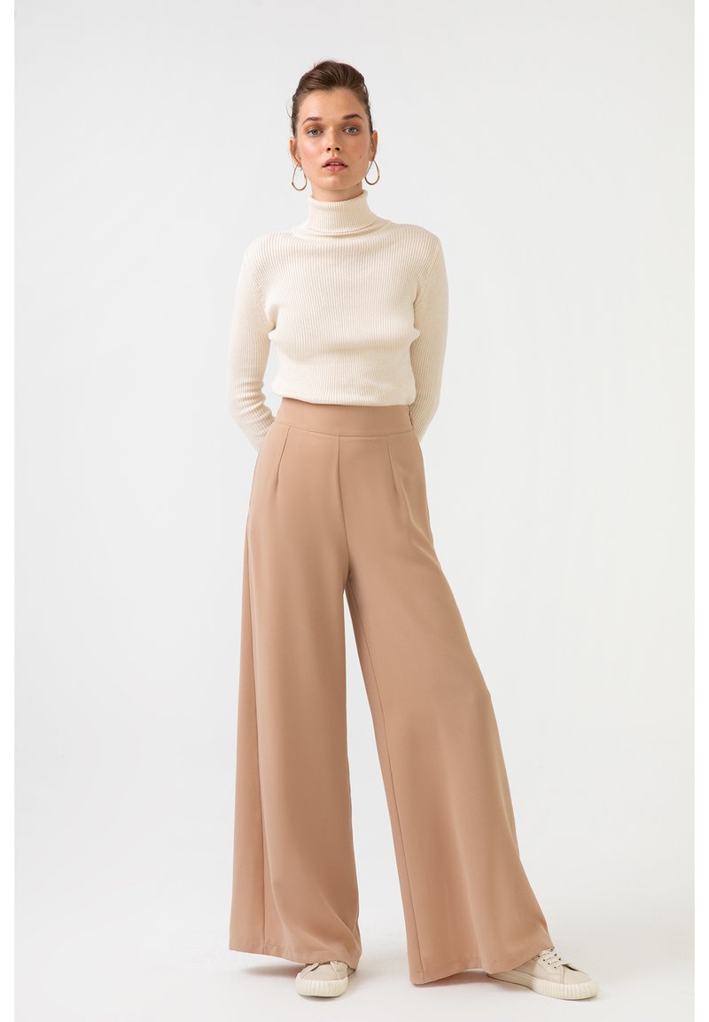 Bež široke hlače z visokim pasom, v kombinaciji s kremno rebrastim puloverjem s turtleneck ovratnikom. Model nosi bele superge. Preprost dizajn.
