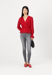 Blusa rossa, strutturata, con maniche lunghe e scollo a V, con vita arricciata, abbinata a jeans skinny grigi e ballerine rosse, mentre tiene una clutch nera.