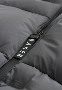 Veste en duvet grise avec une finition lisse et mate. Elle comporte une fermeture éclair noire proéminente et une étiquette en tissu avec le texte "BAKER" en blanc.