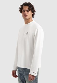 Langarm-T-Shirt aus Baumwolle in Weiß, mit einem schwarzen grafischen Emblem auf der Brust und dem Text "Pure Path" entlang des Ärmels.