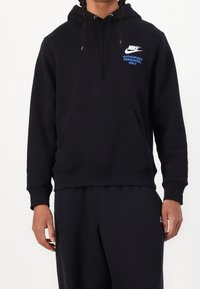 Person klädd i svart Nike-hoodie med framficka och texten "Endast behörig personal" i blått på bröstet, tillsammans med svarta byxor.