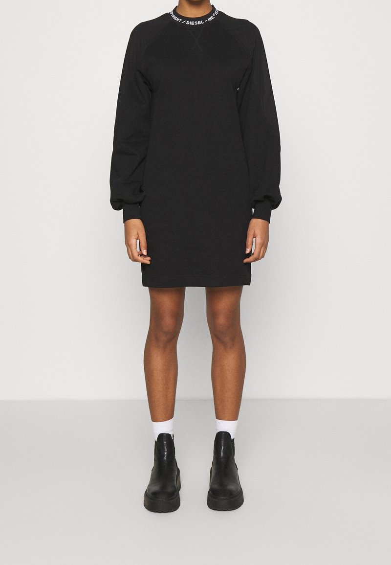 Osoba oblečená v černém svetru šatu s dlouhým rukávem a bílým logem na límci, bílých kotníkových ponožkách a černých robustních kotníkových botách.