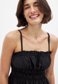 Robe noire sans manches en tissu doux, avec des détails froncés sur le haut et une taille élastique pour une silhouette ajustée.