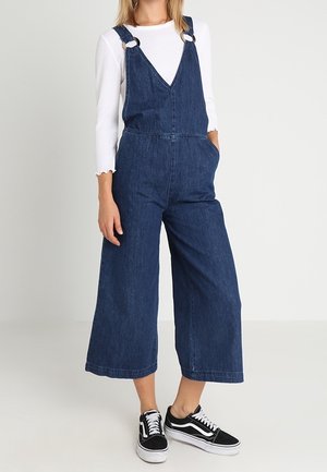 Femme portant une salopette large en denim bleu foncé, un haut à manches longues blanc et des baskets noir et blanc, debout avec une main dans la poche.