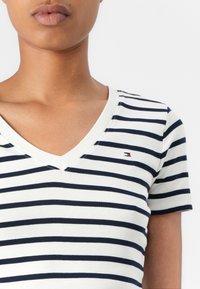 Nærbillede af en person iført en hvid og navyblå stribet V-hals kortærmet skjorte med et lille rektangulært logo på brystet.