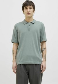 Polo shirt vert clair avec une texture douce, patte de boutonnage à trois boutons et manches courtes. Associé à un pantalon gris ajusté.