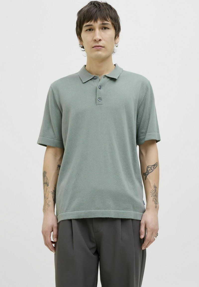 Polo shirt vert clair avec une texture douce, patte de boutonnage à trois boutons et manches courtes. Associé à un pantalon gris ajusté.