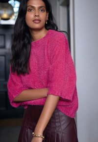 Pūkuotas ryškiai rožinis trumpomis rankovėmis megztinis sluoksniuotas virš bordo spalvos plisijuotos sijono, pasižymintis laisvu siluetu ir švelnia tekstūra. Auksinės apyrankės akcentas.