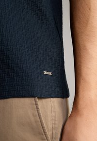 Navyblaues, strukturiertes Strickhemd mit einem gewebten Muster. Ein silbernes Etikett mit "JOOP!" ist am Saum sichtbar. Beigefarbene Hosen sind teilweise sichtbar.