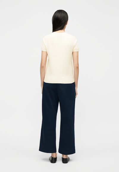 Γυναίκα που στέκεται με την πλάτη στην κάμερα, φορώντας εκρού κοντομάνικη μπλούζα, μπλε σκούρο παντελόνι φαρδύ και μαύρα παπούτσια τύπου slip-on, σε λευκό φόντο.