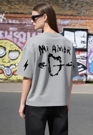 Donna con capelli castani lunghi che indossa occhiali da sole e una maglietta grigia con testo e grafica "Mi Amor", in piedi vicino a un muro di mattoni coperto di graffiti.