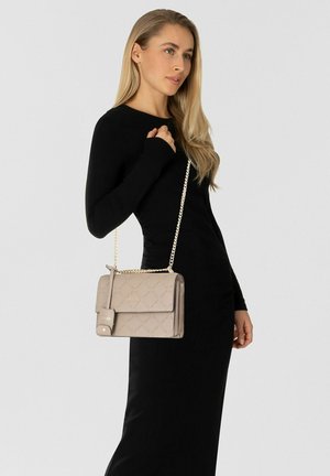 Vrouw in een zwarte lange mouwen jurk draagt een beige gewatteerde handtas met een gouden kettingriem over haar schouder.