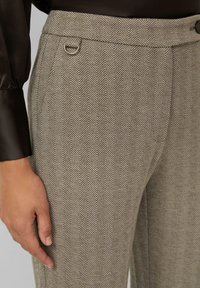 Gros plan sur une personne portant un pantalon beige à motif chevrons avec un détail en anneau métallique et une chemise noire à manches longues.