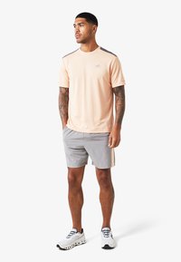 Completo sportivo da uomo composto da una maglietta leggermente pesca traspirante con accenti grigi, abbinata a pantaloni corti grigi e sneakers bianche.