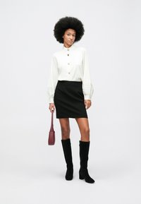 Mulher com cabelo afro, vestindo uma blusa branca de botões, saia preta, botas pretas até ao joelho e segurando uma pequena mala bordô.