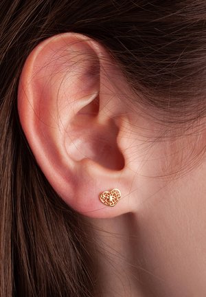 Złote kolczyki w kształcie serca z misternym designem wycięć, umiejscowione blisko ucha, ukazujące gładką teksturę i delikatny styl.