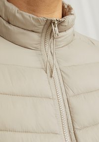 Beige pufferjakke med høj krave, lynlås, quiltet tekstur og let polstring. Designet til varme og komfort.
