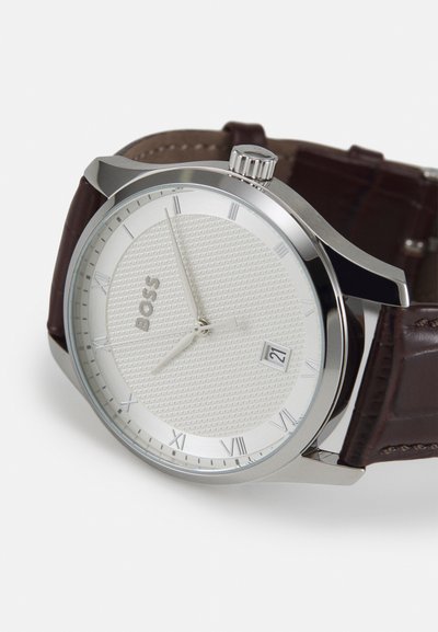 Montre ronde en argent avec un cadran texturé blanc, un bracelet en cuir marron et des chiffres romains comme repères. Dispose d'un affichage de date près de 3 heures.