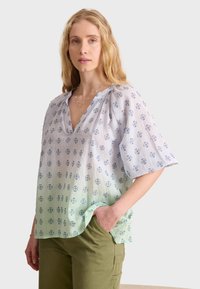 Blusa azul claro con mangas cortas, escote en forma de v y patrones florales en azul marino. Efecto ombre gradual que se desvanece a verde en el dobladillo. Textura suave.