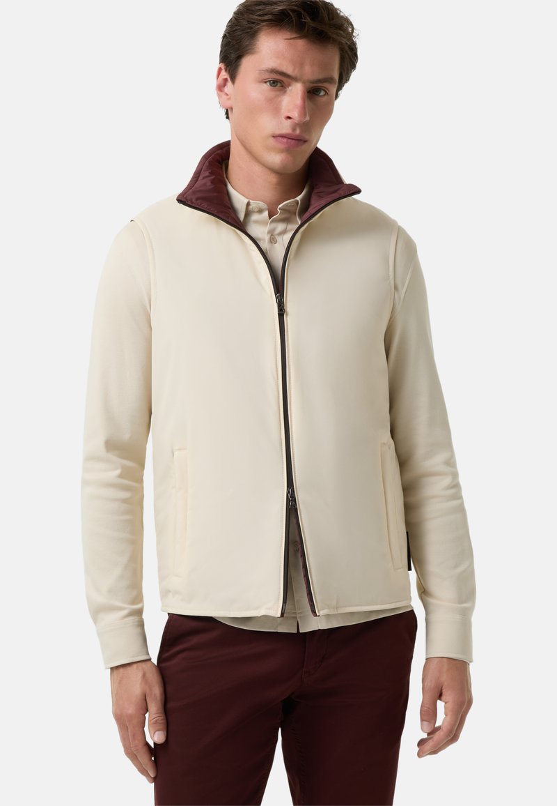 Beige Zip-Up-Jacke mit einem burgunderfarbenen Kragen, Fronttaschen und einer glatten Textur, getragen über einem hellen Hemd und kombiniert mit burgunderfarbenen Hosen.