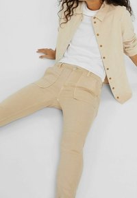 Beżowe dżinsy bawełniane o dopasowanym kroju, z przednimi i tylnymi kieszeniami oraz gładką teksturą, noszone z białą koszulką i jasną kurtką.