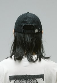 Black denim cap with adjustable strap, rounded crown, and textured fabric, worn backwards over long dark hair, paired with a white t-shirt.