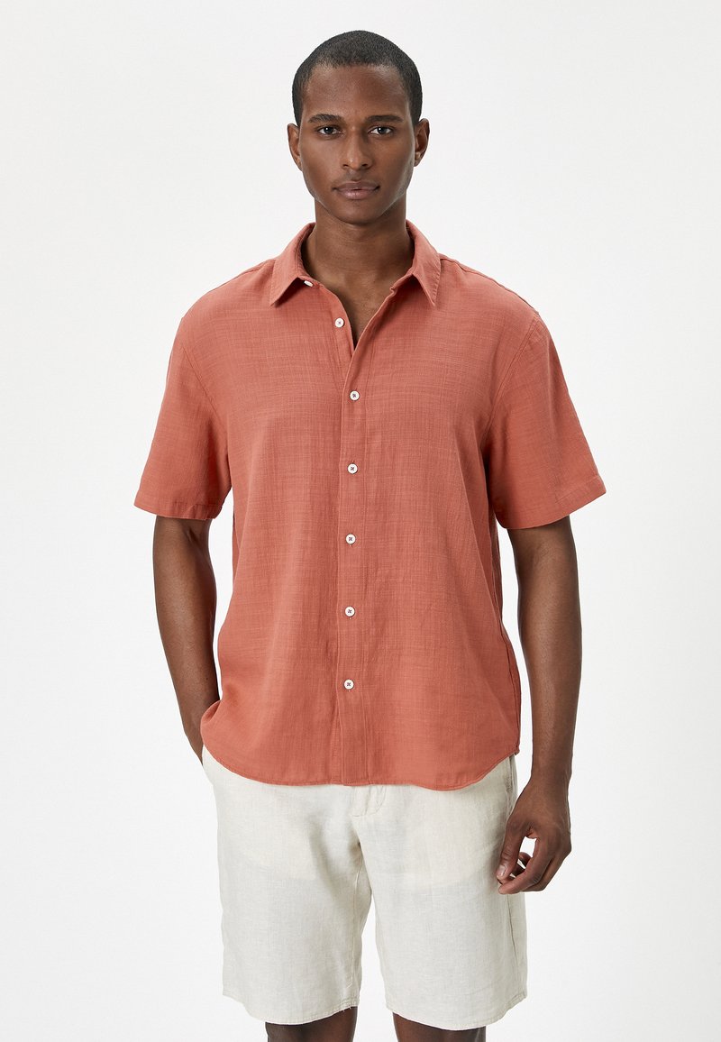 Camisa de manga corta abotonada en lino color naranja quemado con un corte relajado, que presenta un cuello y botones blancos, combinada con pantalones cortos de color beige claro.