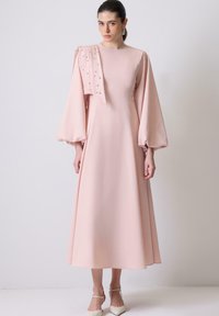 Naisso wearing a pikk heleroosa kleit, millel on puhvärvlid, kaunistatud rippuv õlavarre osa ja valged teravatipulised kõrged kontsad, seistes valge seina vastas.