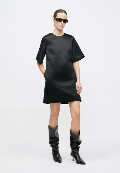 Robe courte en satin noir avec un col rond et des manches courtes, associée à des bottes noires souples montantes jusqu'aux genoux avec des bouts pointus.