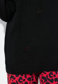Schwarzer Strickpullover mit lockerer Passform, der verwitterte Details aufweist. Kombiniert mit roten Leggings im Leopardenmuster, die einen kräftigen Farbkontrast zeigen.