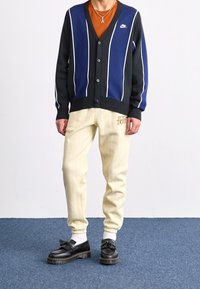 Mustavalkoiset raidalliset neuletakki, oranssi T-paita alla, kermanväriset raidoitetut housut ja mustat liukukengät, joissa on kuvioitu pohja.