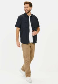 Hemd mit kurzen Ärmeln in Marineblau und zwei Brusttaschen, kombiniert mit beigen Hosen und weißen Sneakers. Das Modell steht vor einem schlichten Hintergrund.