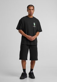 Camiseta negra de gran tamaño con texto gráfico verde, combinada con pantalones cortos negros hasta la rodilla y zapatillas negras, con un tejido suave y un corte relajado.