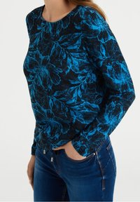 Blusa de manga larga en negro con un patrón floral azul. La tela es texturizada, presenta un escote redondo y detalles laterales con herrajes plateados.