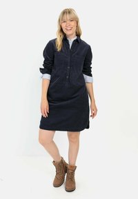 Granatowa sukienka z korduroyu z zapięciem na guziki z przodu, dwoma kieszeniami na piersi, podwiniętymi rękawami i lekko rozszerzanym dołem. Połączona z brązowymi butami trekkingowymi.