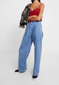 Jeans ampi a vita alta di colore azzurro chiaro, con pieghe frontali, abbinati a un top in satin rosso e a una giacca camouflage. Sono indossate sneakers nere.