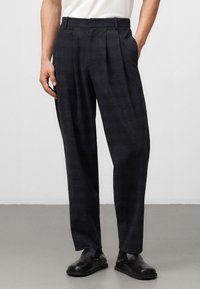 Donker geruite broeken van gestructureerde stof, met een hoge taille, plooien en zijzakken; gedragen met zwarte instapschoenen.