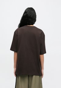 Άτομο με μακριά μαύρα μαλλιά, που φοράει φαρδιά σκουρόχρωμη καφέ μπλούζα και παντελόνι χακί, στέκεται με το πρόσωπο προς ένα απλό ανοιχτόχρωμο φόντο.