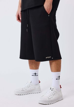 Homme portant un short noir au genou, des chaussettes blanches avec des logos noirs et des baskets blanches, debout devant un fond uni.
