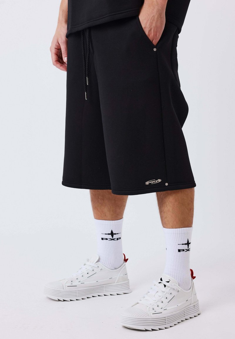 Homme portant un short noir au genou, des chaussettes blanches avec des logos noirs et des baskets blanches, debout devant un fond uni.